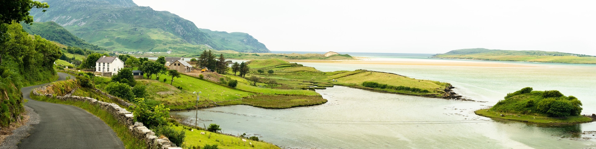 Un paisaje costero con una carretera sinuosa, una casa, campos verdes, un río y montañas lejanas.