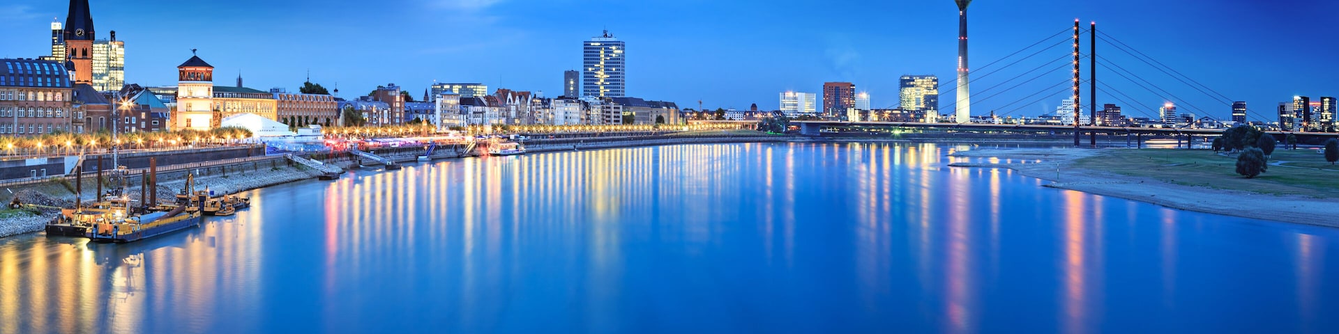 Horizonte urbano ao entardecer, com um rio refletindo luzes, uma torre imponente e uma ponte.