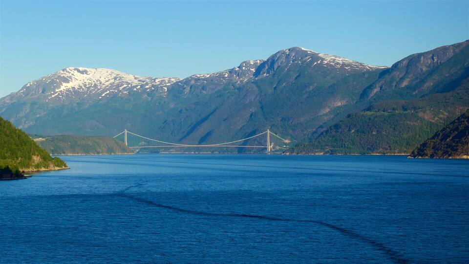 A suspension bridge over a calm body of water, surrounded by mountains with snow-capped peaks.