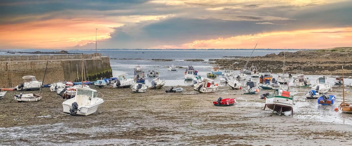 A coastal scene with numerous boats anchored on a muddy shore, a rocky outcrop, and a distant city skyline under a colorful sunset.