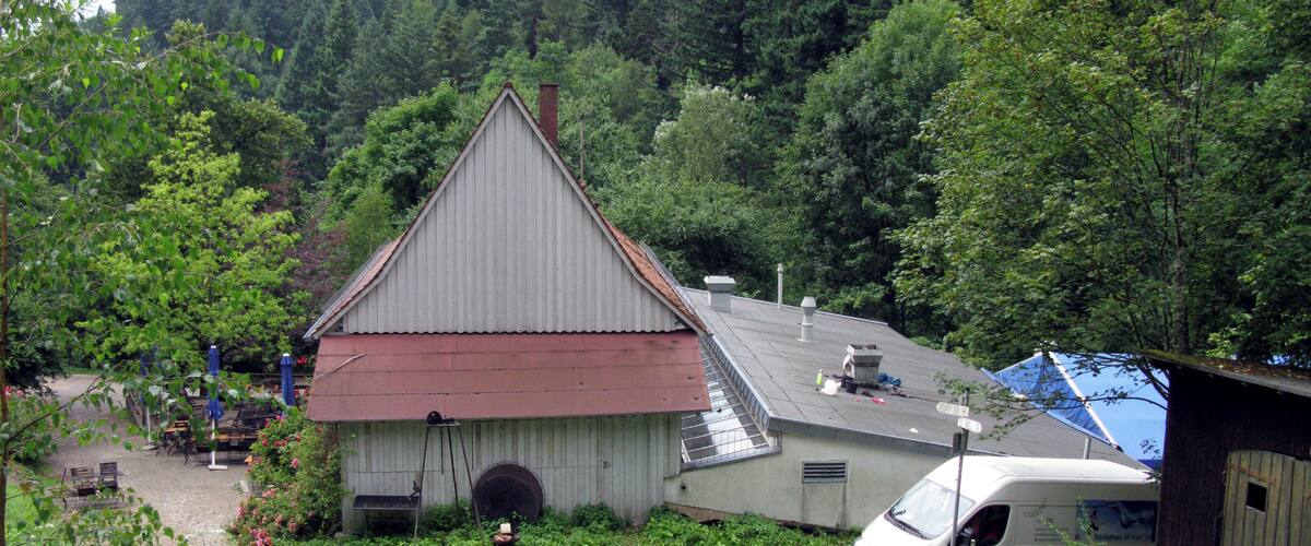 A rustic building with a red roof and a chimney, surrounded by lush greenery and a forested hillside.