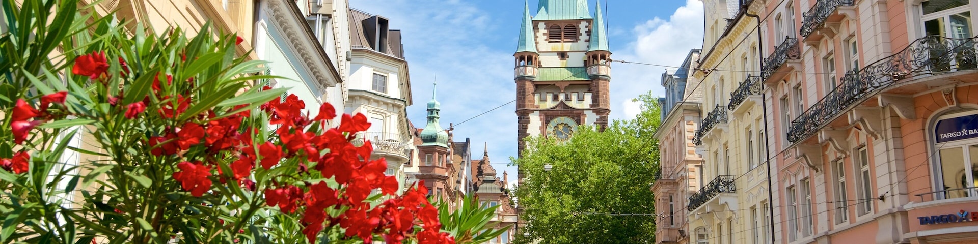 Rua com edifícios históricos, torre de igreja e flores vermelhas em primeiro plano.