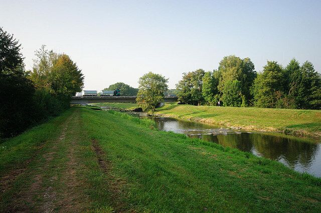 A grassy path beside a calm river, trees, and a distant bridge.