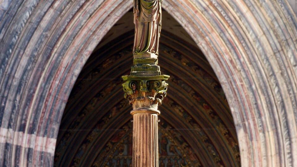 A Gothic-style stone column with a statue at the top, set against a large arched doorway with intricate stone carvings.