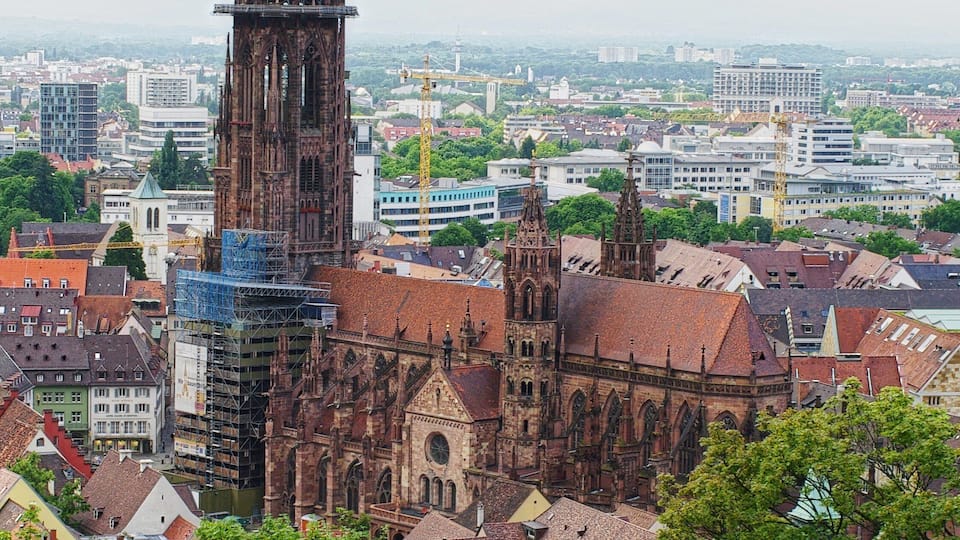 A historic cathedral with a tall spire and intricate architecture, surrounded by modern buildings and greenery.
