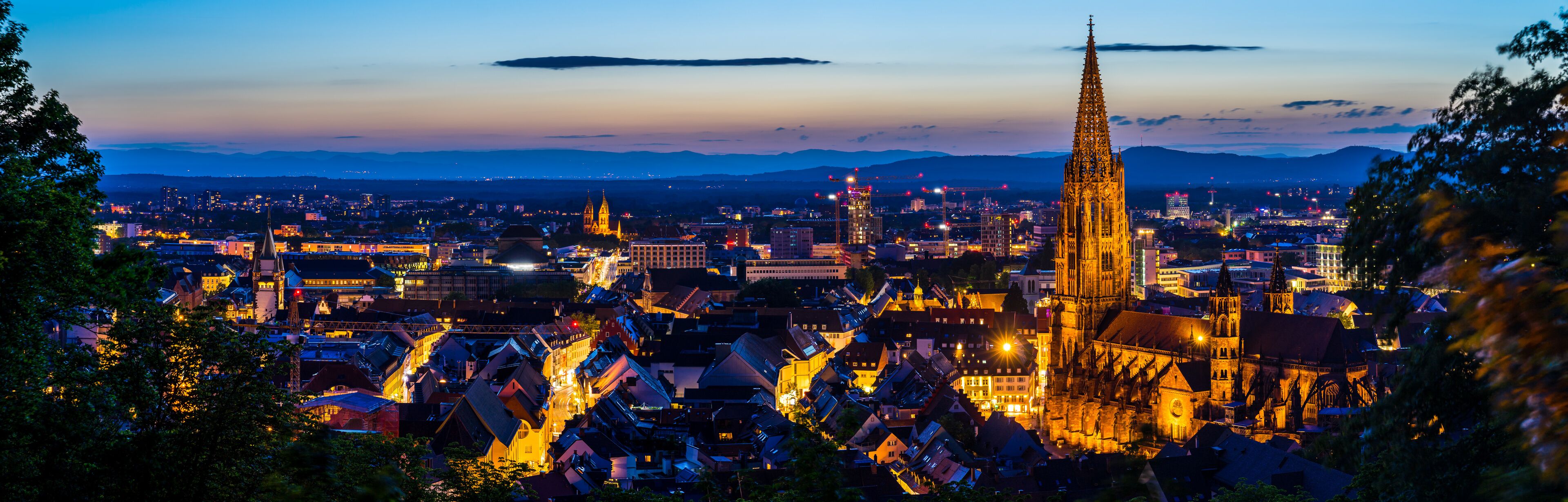 Paisagem urbana ao anoitecer com edifícios iluminados, uma catedral imponente e montanhas ao fundo.
