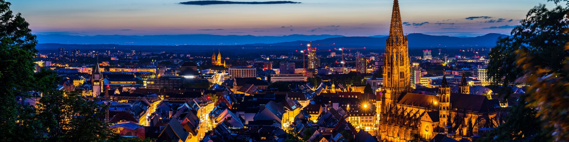 Paisagem urbana ao anoitecer com edifícios iluminados, uma catedral imponente e montanhas ao fundo.