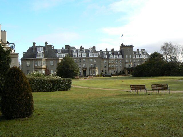 A large, historic building with a well-maintained lawn and benches in the foreground.
