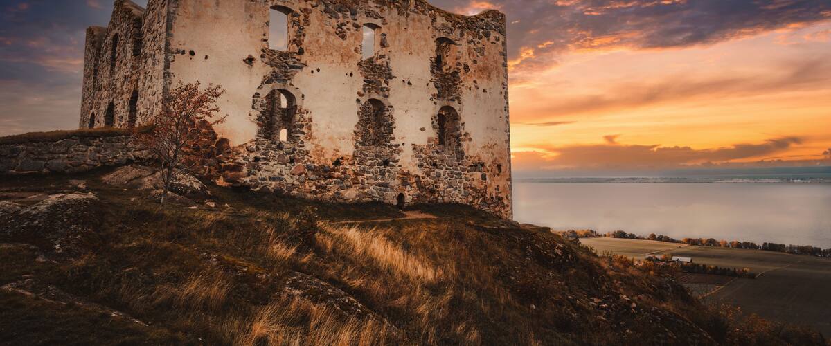 An ancient stone building with arched windows, surrounded by grassy terrain and overlooking a body of water.