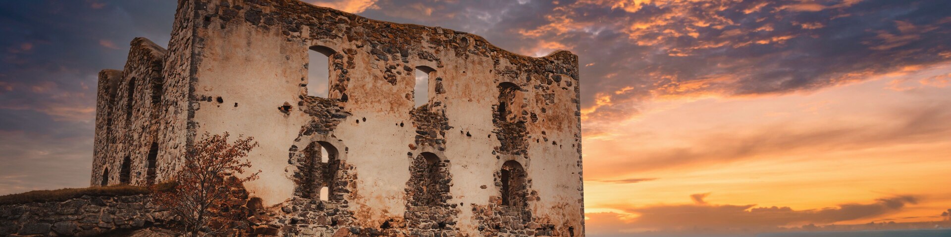 An ancient stone building with arched windows, surrounded by grassy terrain and overlooking a body of water.