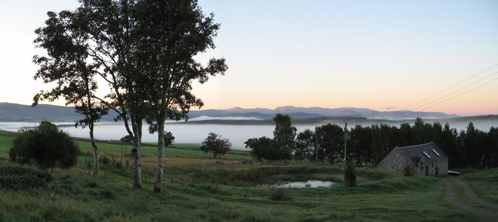 A rural landscape with a large tree, a stone building, and a body of water in the distance.