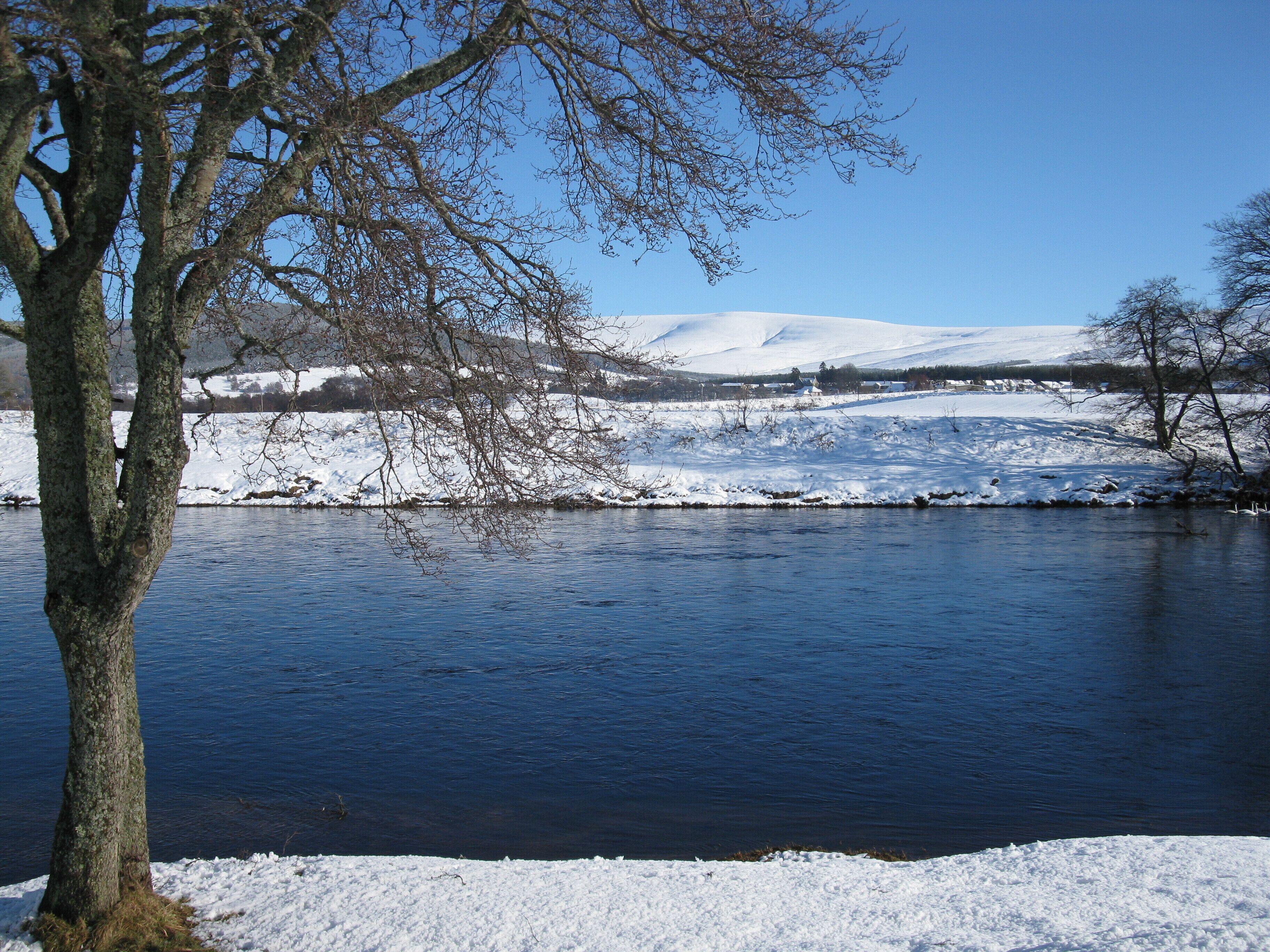 A snow-covered landscape with a river, trees, and distant hills.