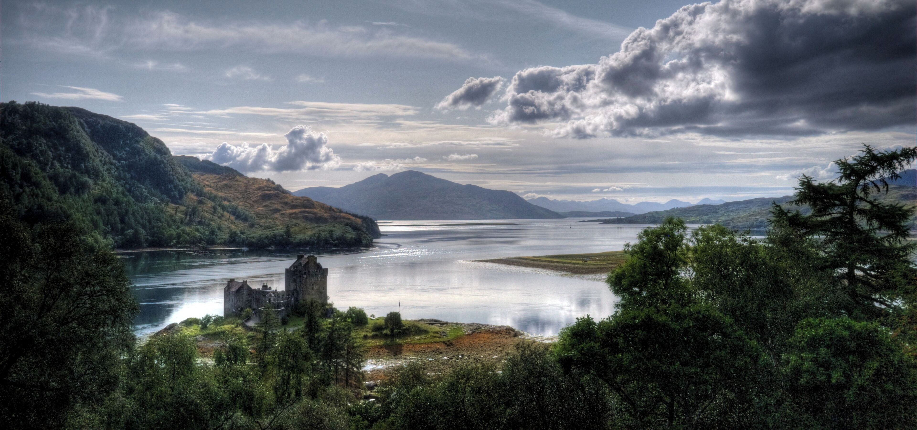 A historic castle by a calm lake surrounded by lush green hills and mountains under a cloudy sky.