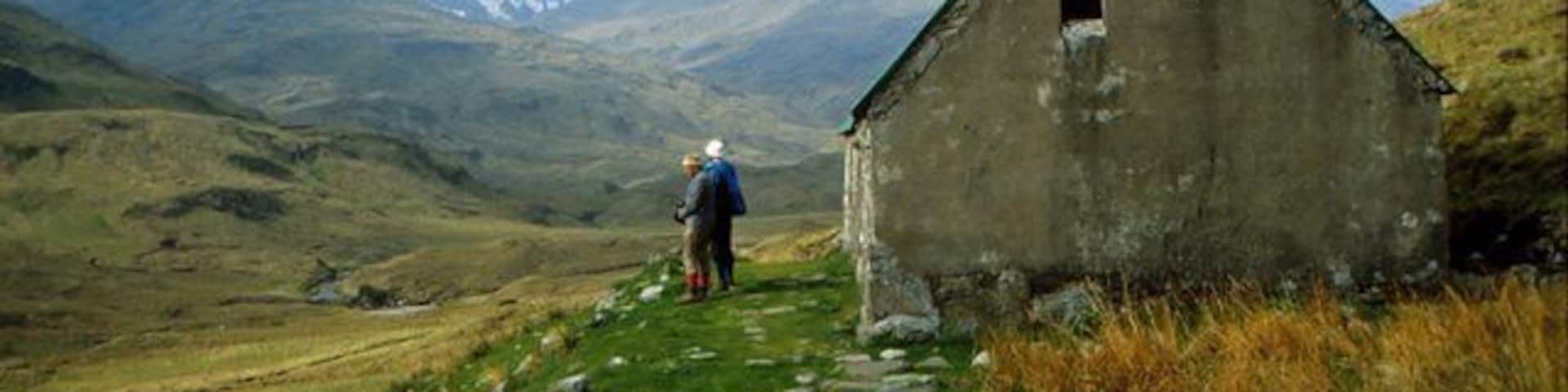 A person walking towards a small stone building in a grassy valley with mountains in the background.