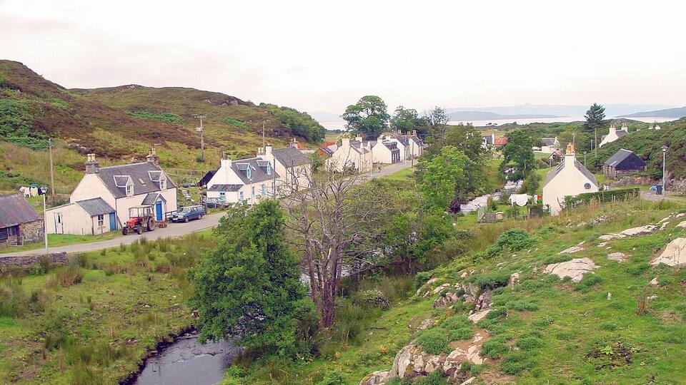 A rural village with houses, a narrow stream, and rolling hills in the background.