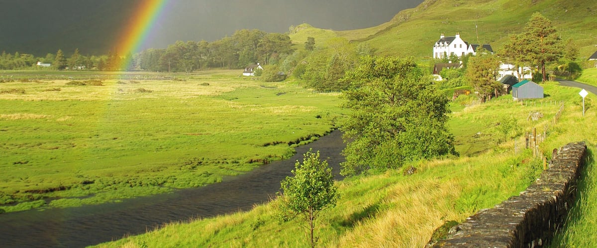 A rainbow over a rural landscape with a stone wall, a river, and houses in the distance.