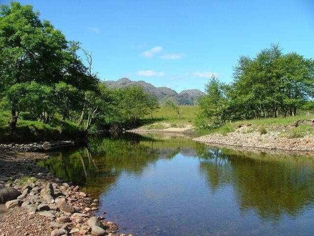 Un río tranquilo con orillas rocosas, rodeado de frondosos árboles verdes y montañas distantes.