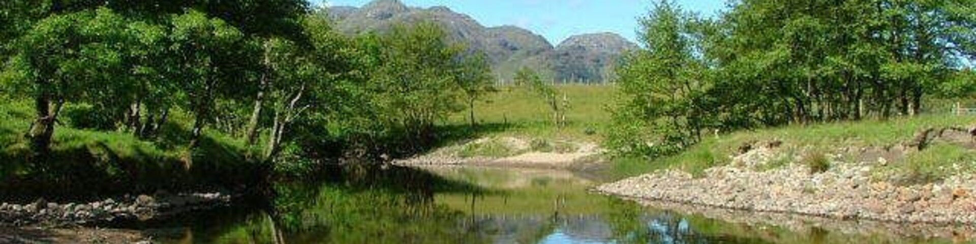 Un río tranquilo con orillas rocosas, rodeado de frondosos árboles verdes y montañas distantes.