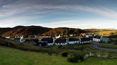 A coastal village with white houses, a winding road, and rolling hills in the background.