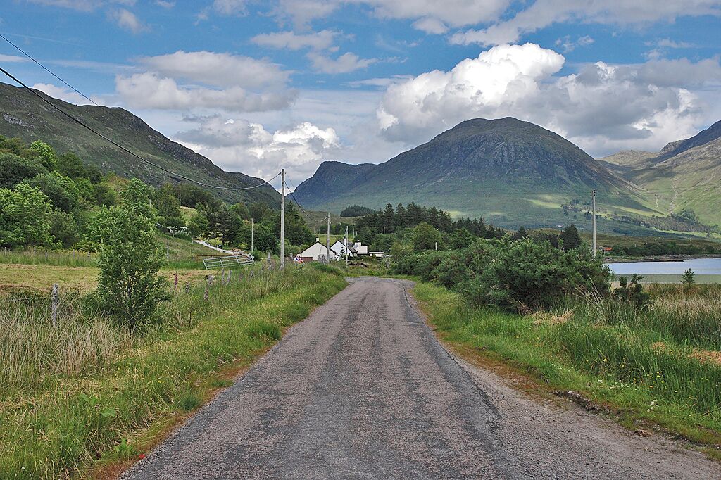 A rural road flanked by lush greenery and mountains.