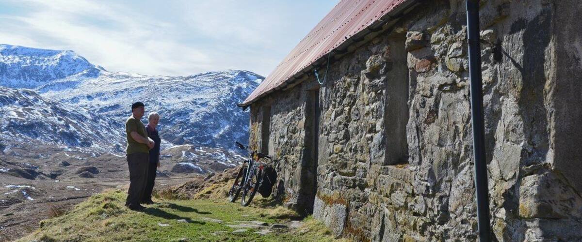 A stone building with a red roof, a mountain range in the background, and a person standing in the foreground.