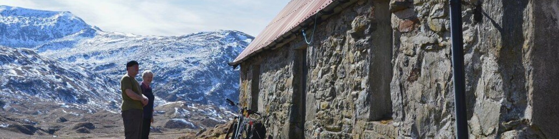 A stone building with a red roof, a mountain range in the background, and a person standing in the foreground.
