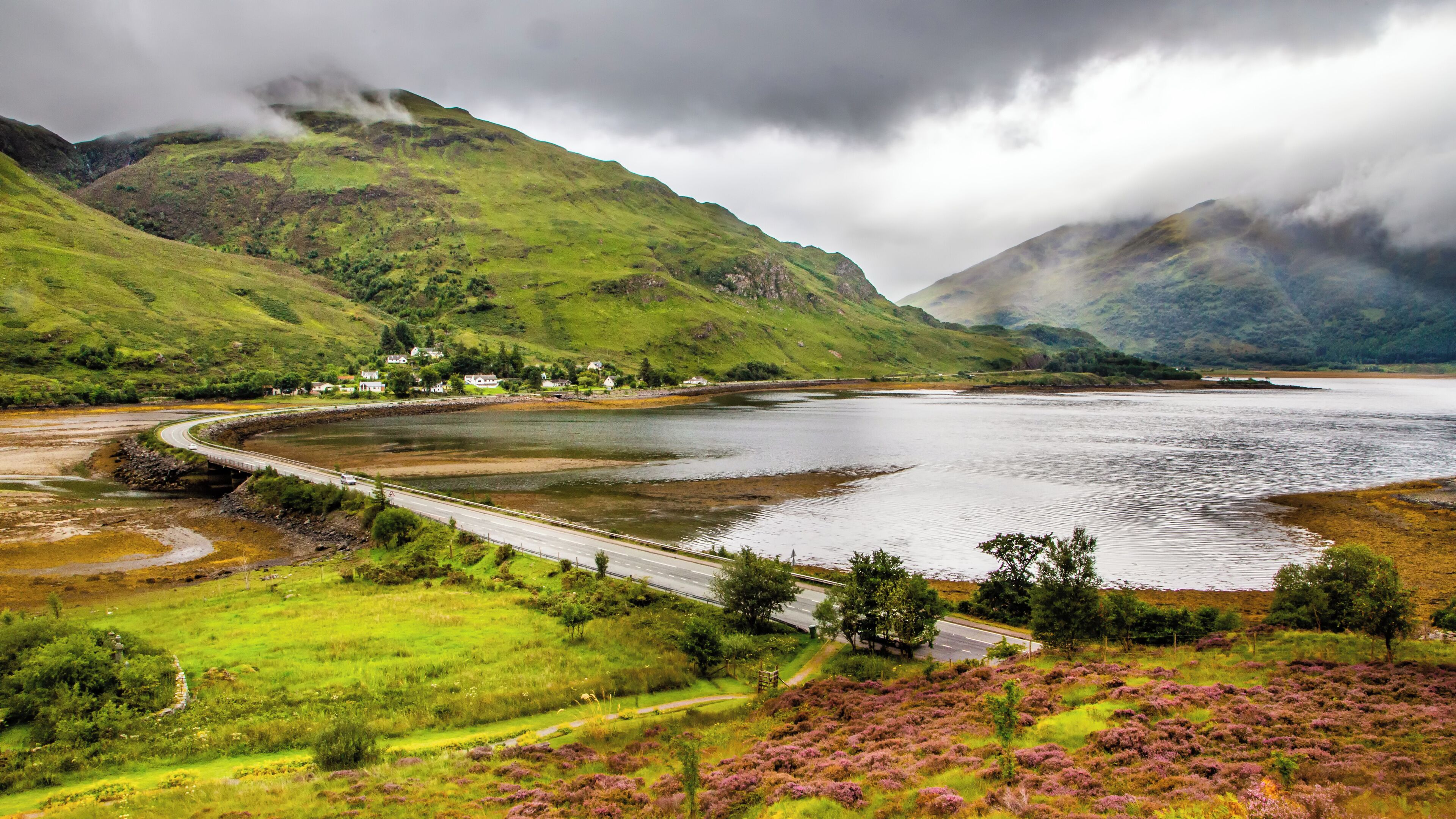 A coastal landscape with a road curving around a bay, houses on a hillside, and heather-covered hills.