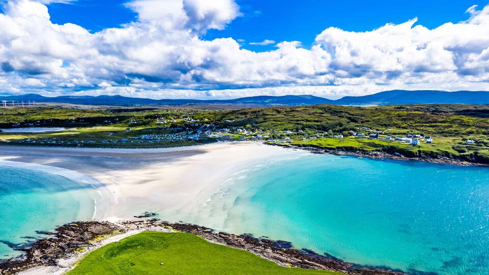 A coastal landscape with a sandy beach, turquoise waters, and a green hillside with scattered buildings.
