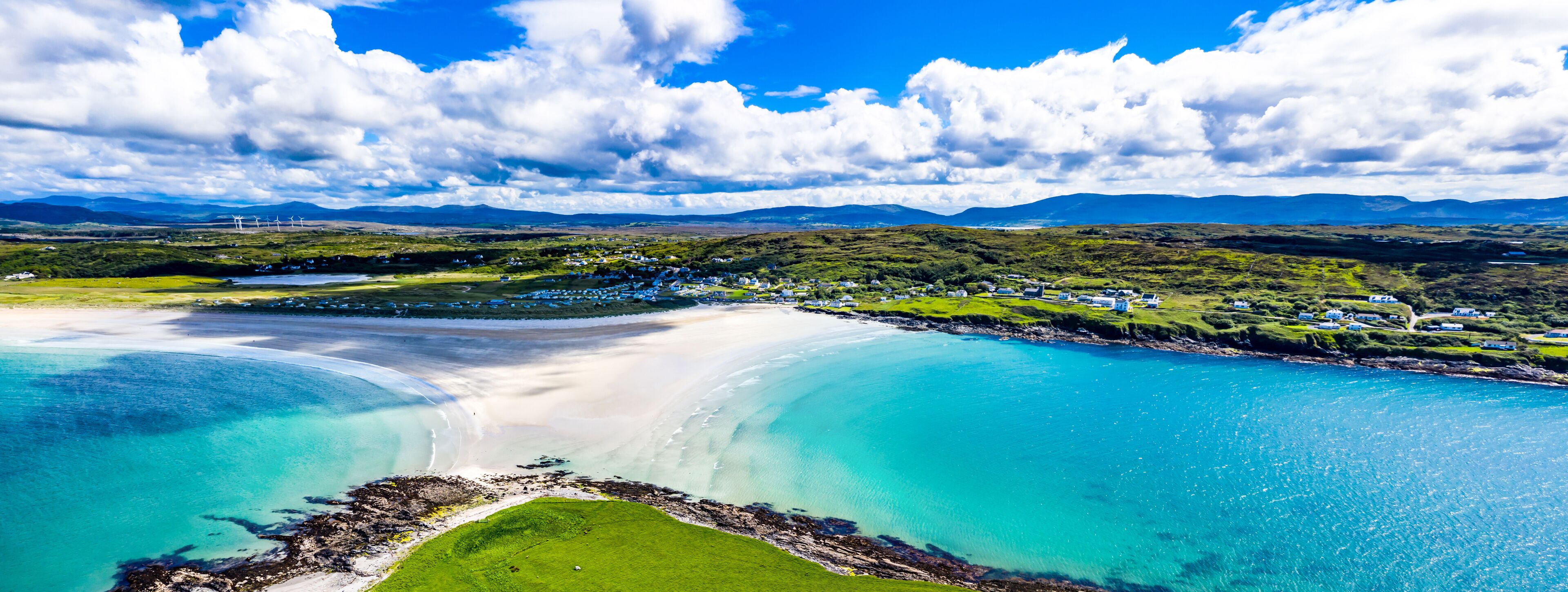 A coastal landscape with a sandy beach, turquoise waters, and a green hillside with scattered buildings.