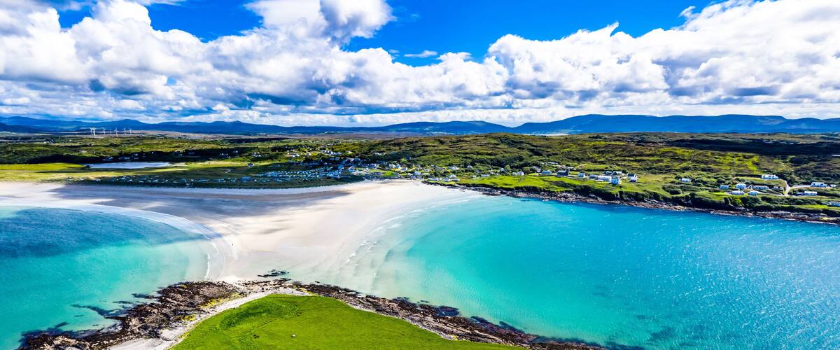 A coastal landscape with a sandy beach, turquoise waters, and a green hillside with scattered buildings.