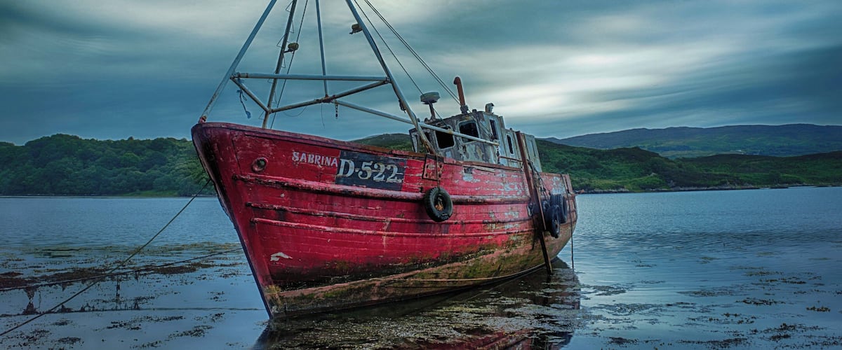 A red fishing boat named 'Sabrina D 522' anchored in calm waters with a backdrop of green hills under a cloudy sky.