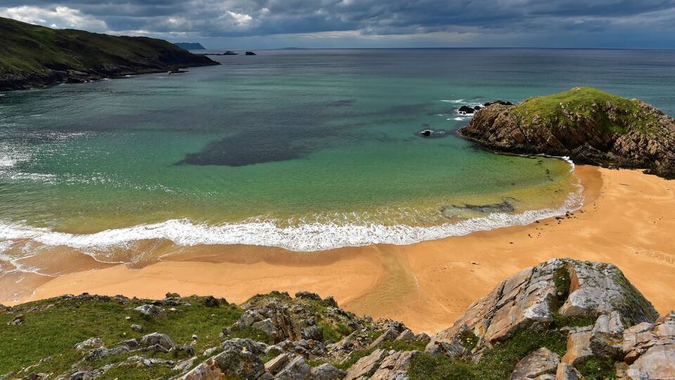 A coastal landscape with a sandy beach, rocky outcrops, and a grassy hill.