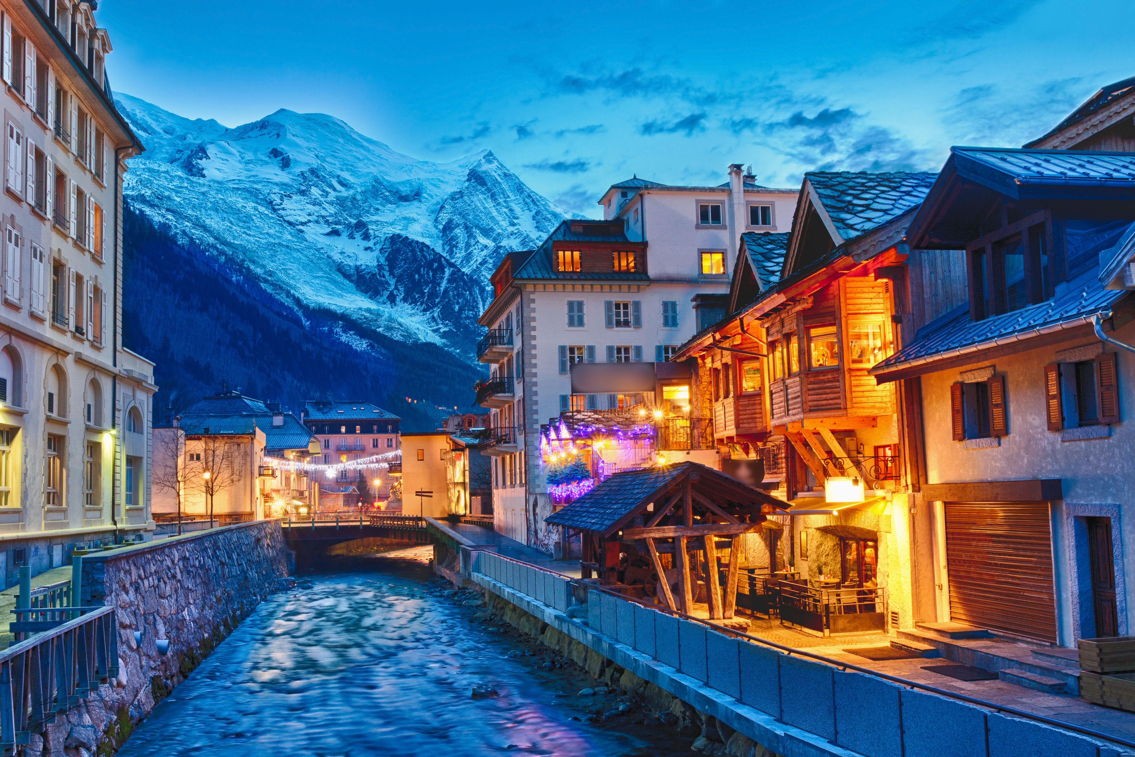 A snow-covered mountain range in the background, a river running through a town with buildings on either side, and a wooden bridge in the foreground.