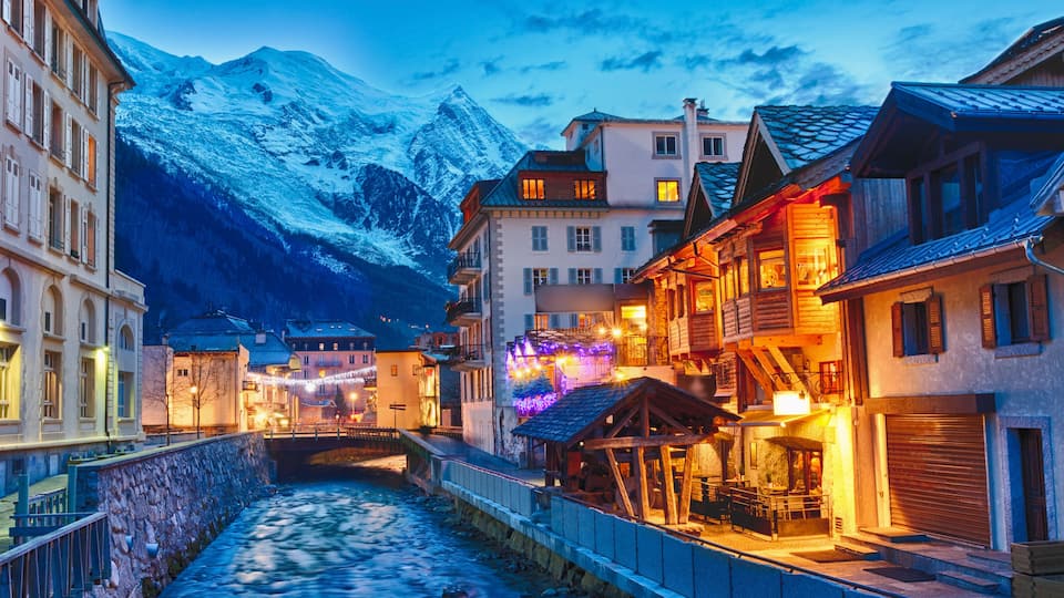 A snow-covered mountain range in the background, a river running through a town with buildings on either side, and a wooden bridge in the foreground.