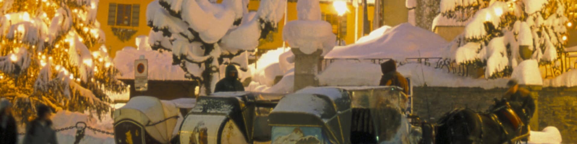 A snow-covered street with vintage cars and a horse-drawn carriage.