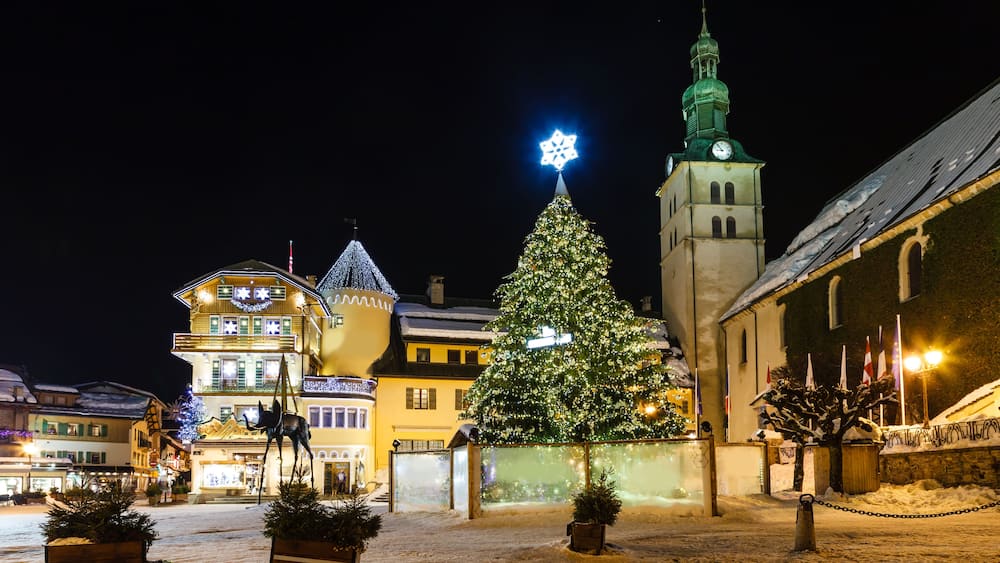 Ein verschneiter Platz mit einem geschmückten Weihnachtsbaum, einer Kirche mit Kirchturm und traditionellen Gebäuden mit Lichterketten.