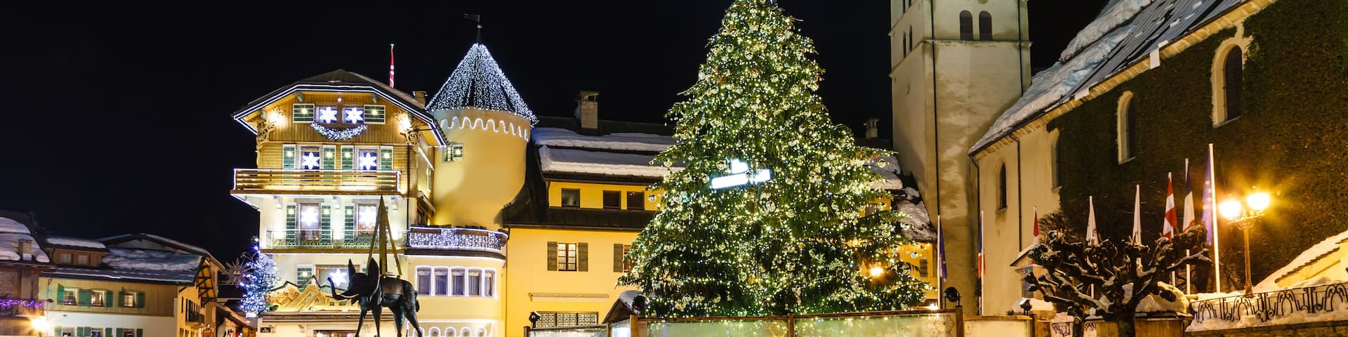 A snow-covered square with a decorated Christmas tree, a church with a spire, and traditional buildings with lights.