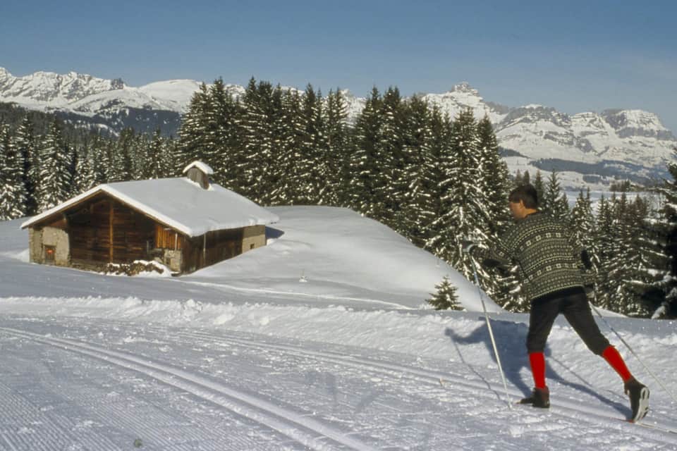 Een persoon skiënd op een besneeuwde helling, met een houten chalet en besneeuwde bomen op de achtergrond.