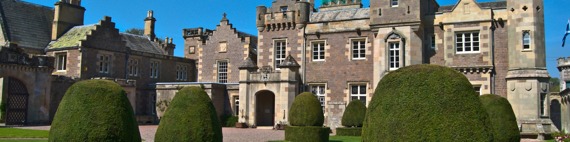 A historic building with a tower, surrounded by well-manicured hedges and a gravel path.
