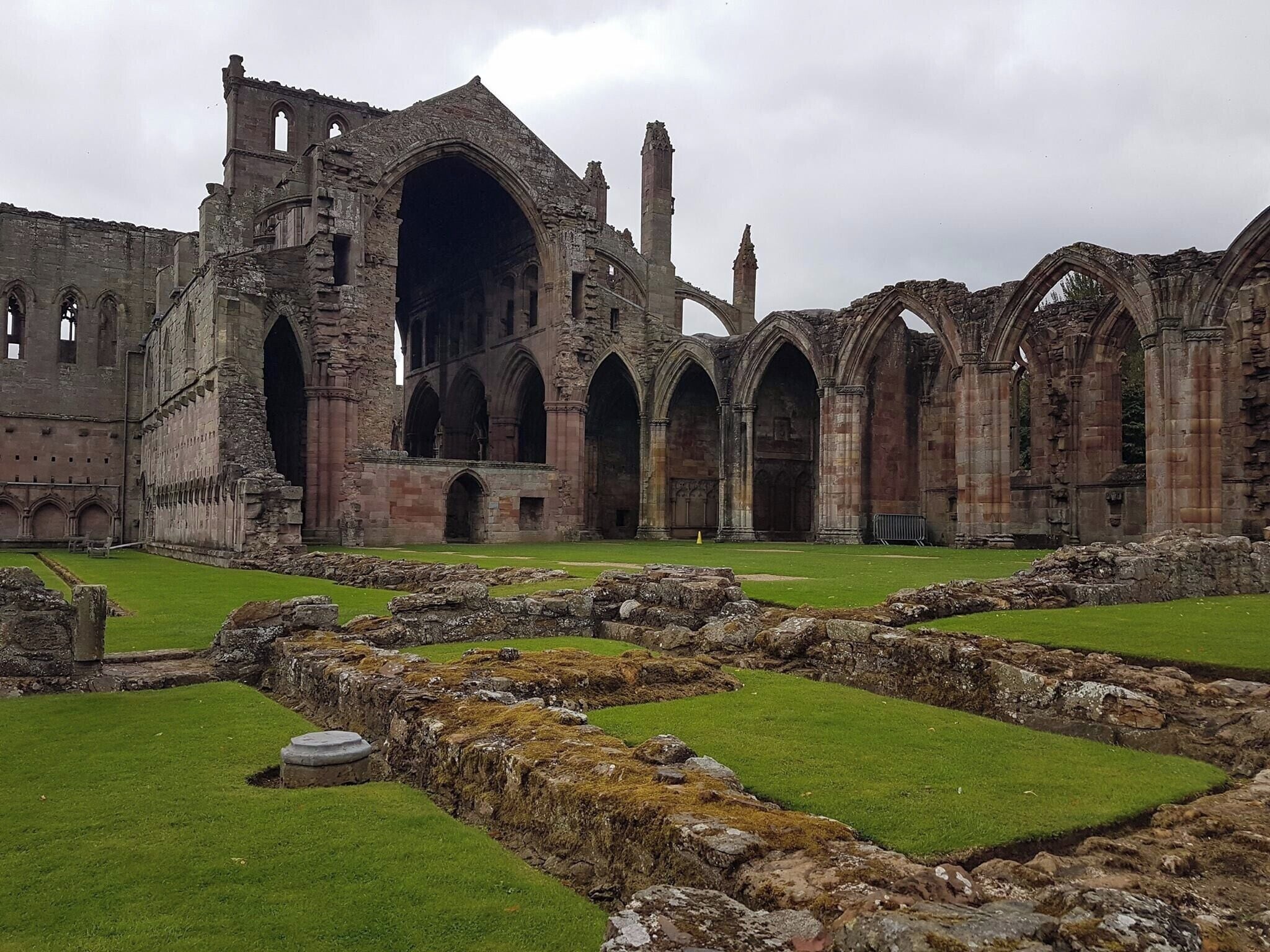 The ruins of a large stone building with multiple arches and columns, surrounded by green grass and scattered stones.