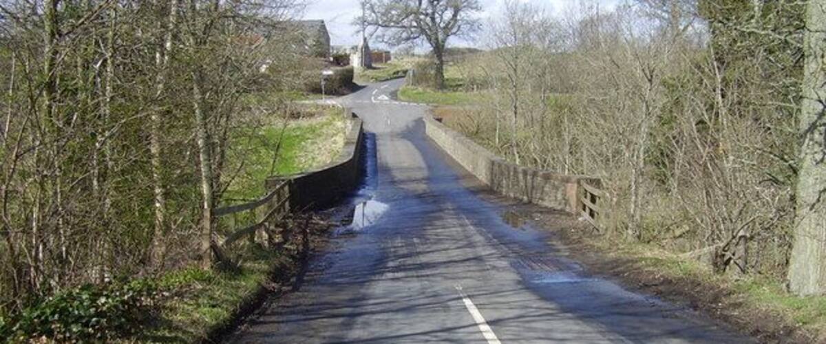 A rural road flanked by trees and a stone wall, leading to a distant building.