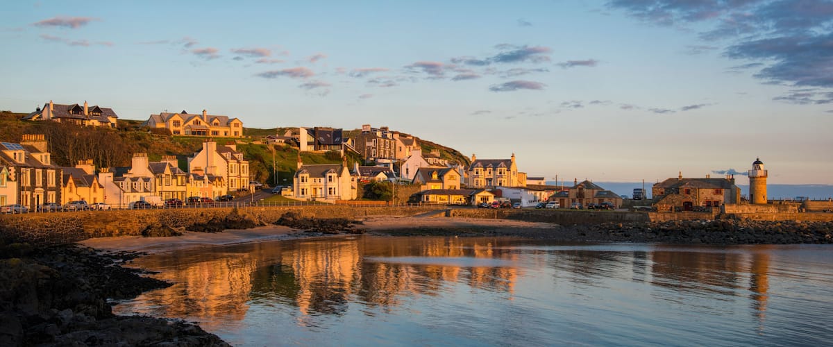 A coastal town with historic buildings, a lighthouse, and calm waters reflecting the structures.