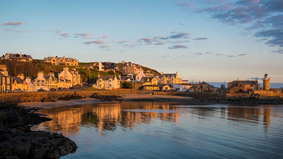A coastal town with historic buildings, a lighthouse, and calm waters reflecting the structures.