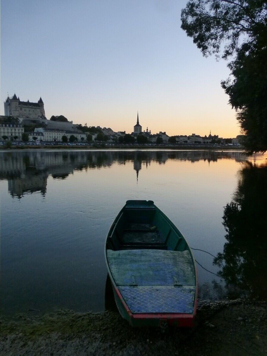 A calm river with a small boat docked on the shore, a castle in the background, and a church spire.