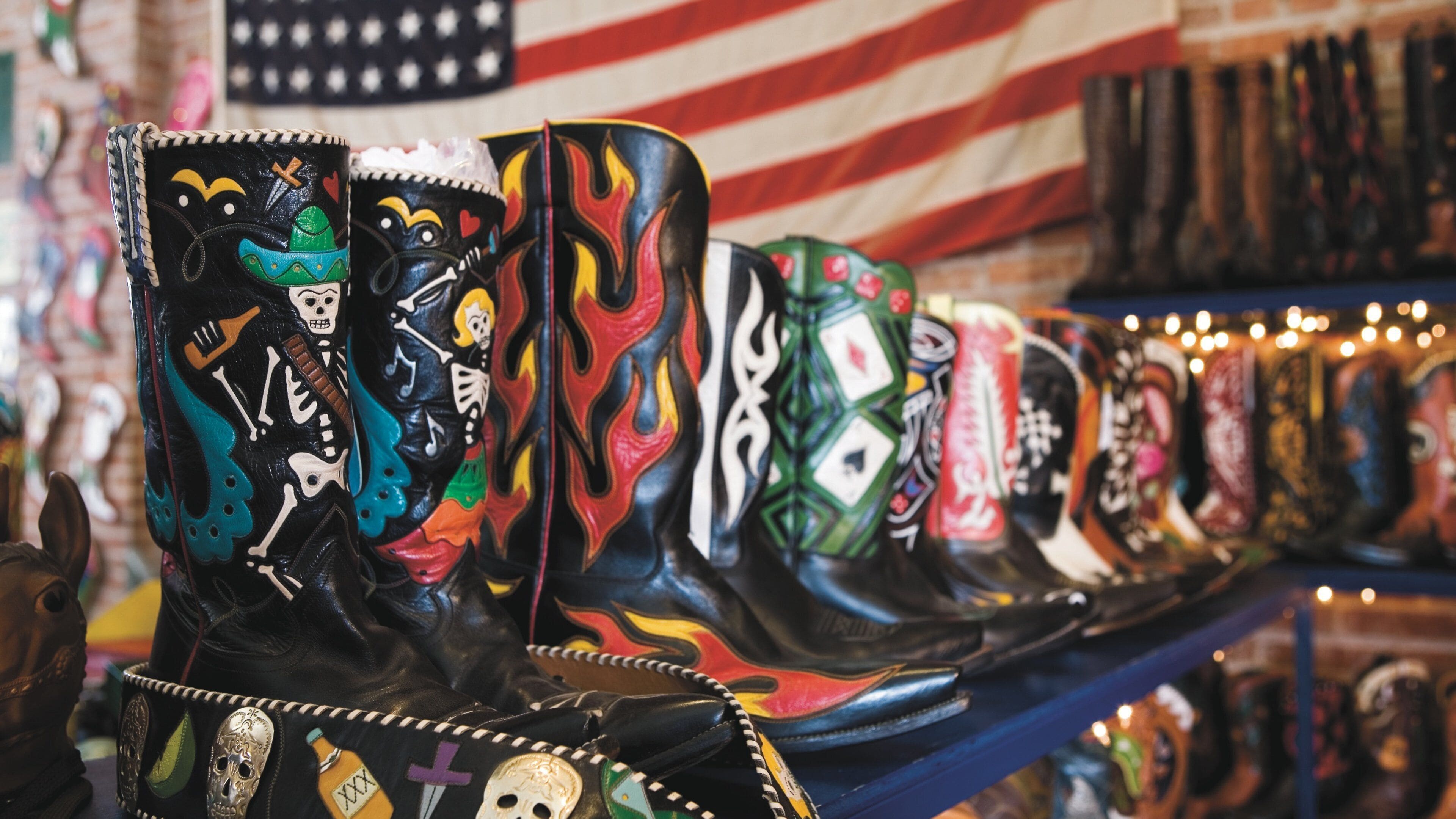 Exposição de botas coloridas com vários desenhos, incluindo caveiras e chamas, em frente a uma bandeira americana.