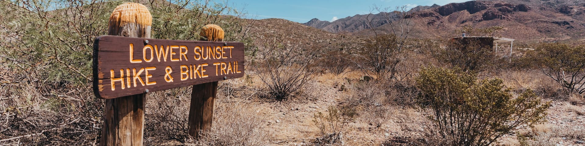 A wooden signpost with 'Lower Sunset Hike & Bike Trail' written on it, set against a desert landscape with sparse vegetation and distant mountains.