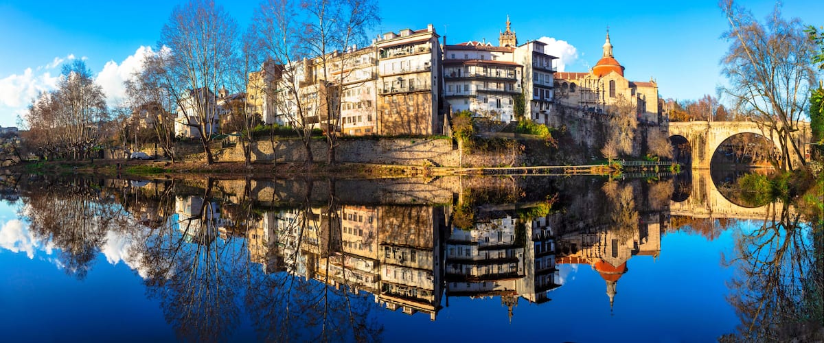 A historic cityscape with a river reflecting buildings, a prominent church with a dome, and a stone bridge.