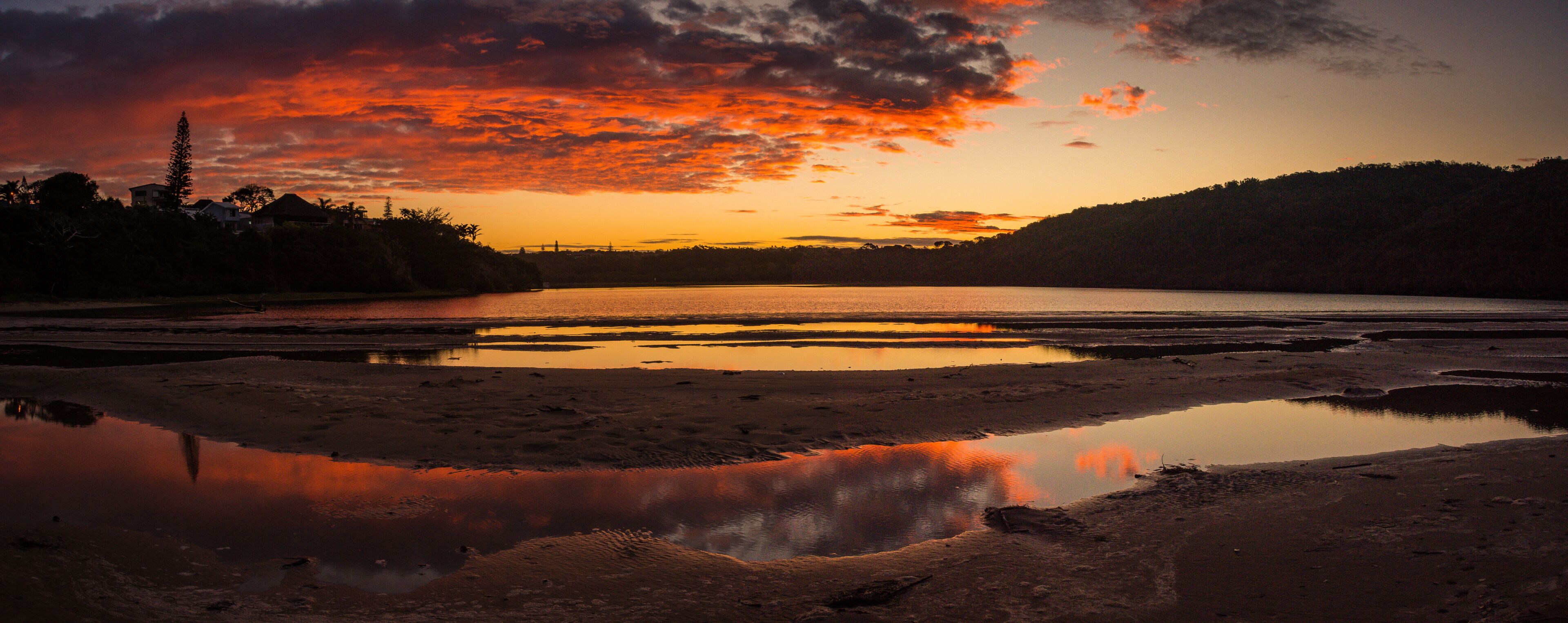 Um pôr do sol sobre um lago calmo com reflexos, cercado por colinas e vegetação escassa.