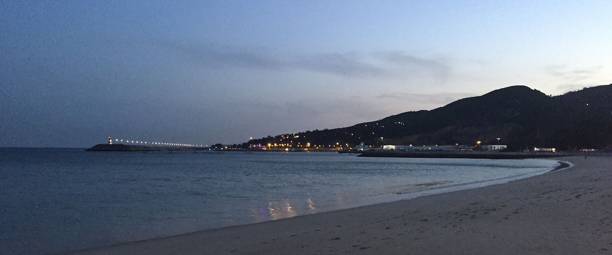 A beach with footprints leading to the water, a distant shoreline with lights, and a mountain in the background.