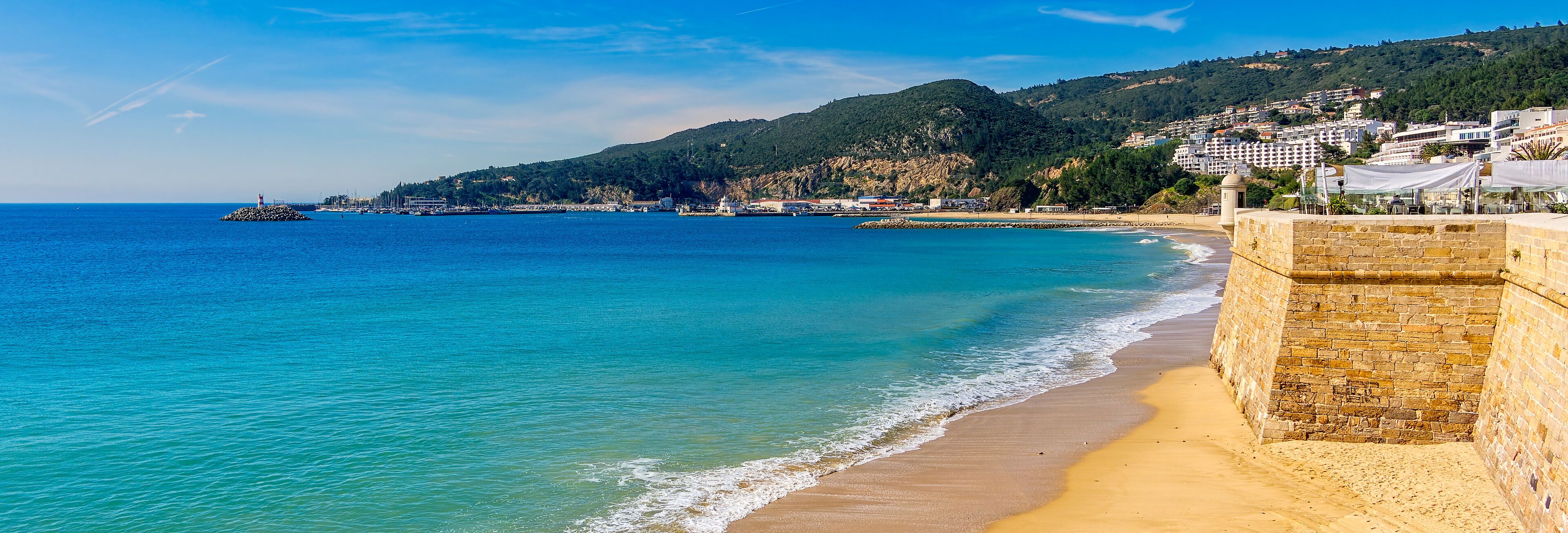 A coastal scene with a sandy beach, a stone wall, and a backdrop of hills and buildings.